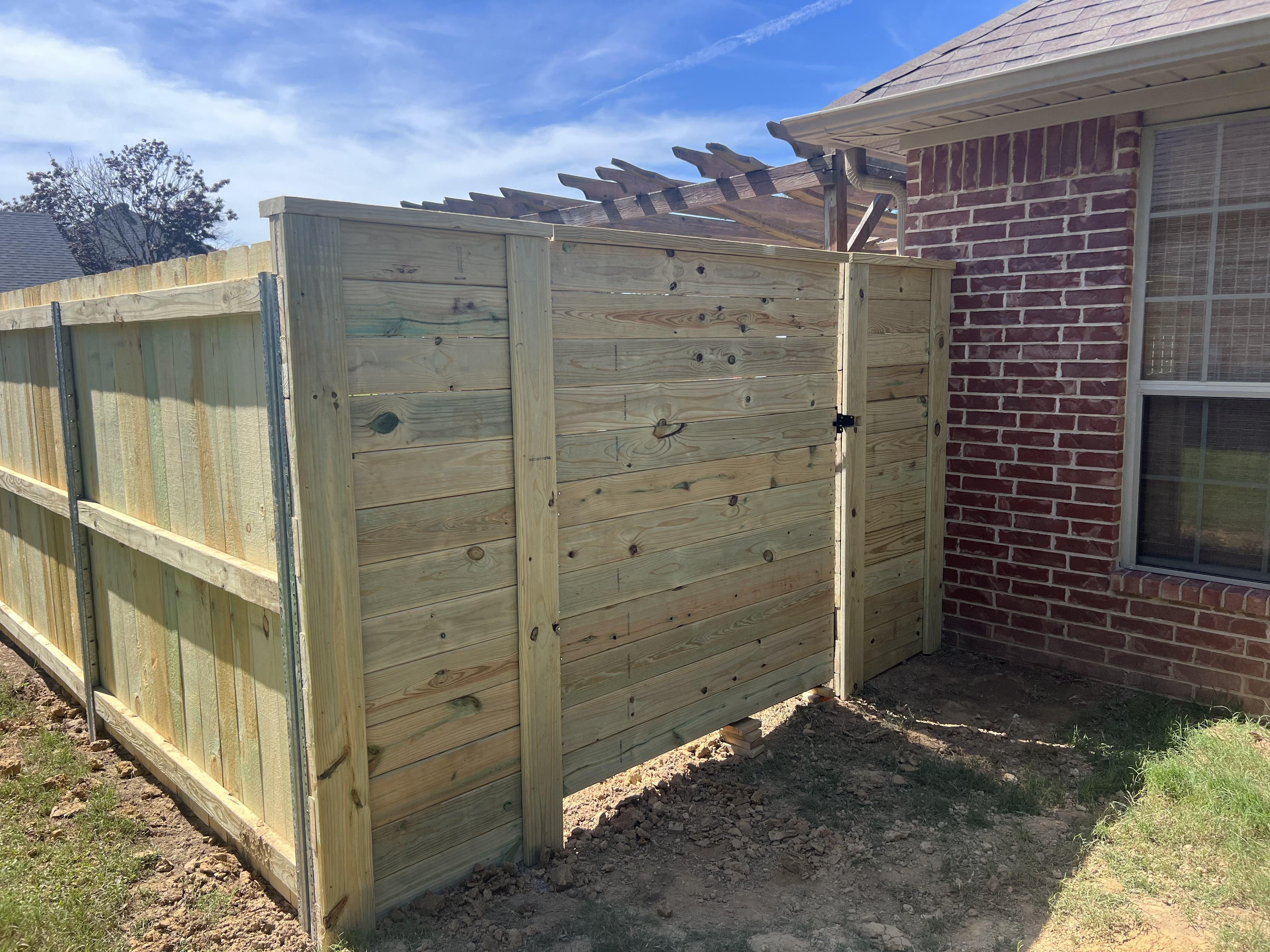 Horizontal-board wood screen with a walk gate built against a brick house under a wood pergola, with a vertical board fence on steel posts continuing to the left